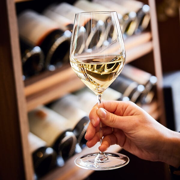 A hand holding a glass of white wine in front of a wine rack filled with bottles.
