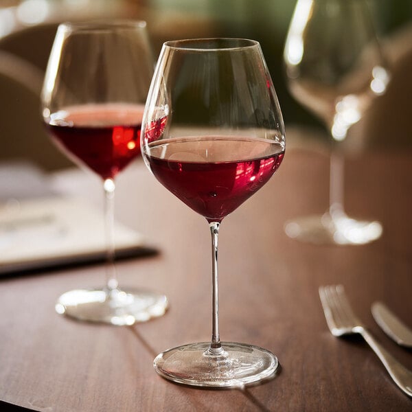 A clear wine glass filled with red wine is placed on a wooden table, with additional wine glasses and a fork visible in the background.