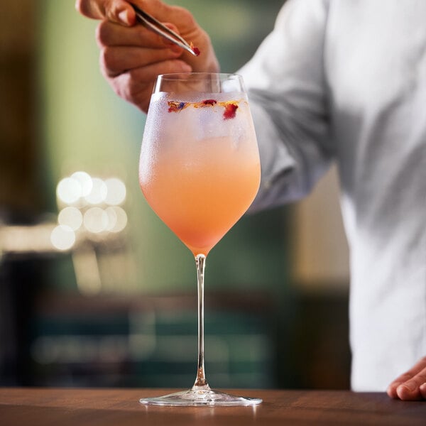 A person is garnishing a stemmed glass filled with a light orange-pink beverage and ice.