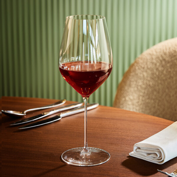 A clear wine glass partially filled with red wine is placed on a wooden table next to a folded white napkin and silverware.