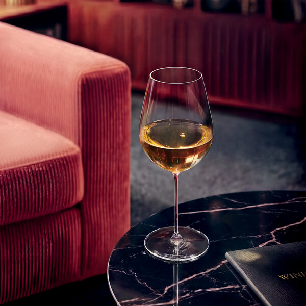 A wine glass filled with white wine is placed on a round black marble table next to a pink upholstered sofa.