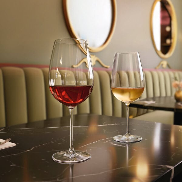 Two wine glasses, one filled with red wine and the other with white wine, are placed on a dark marble table in a stylish restaurant setting with upholstered seating and round mirrors on the wall.