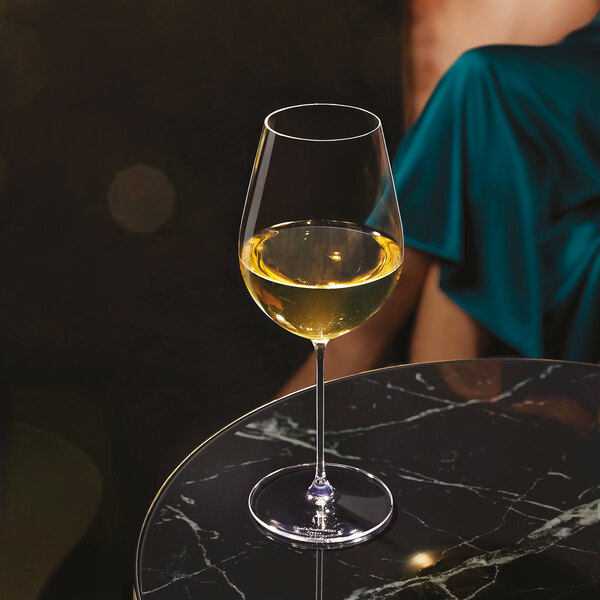 A clear wine glass filled with white wine is placed on a round, black marble table, with a person in a teal dress sitting in the background.