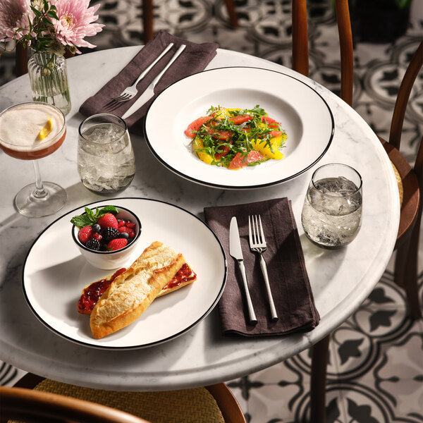 A round white plate with a thin black band around the rim, holding a sandwich with jam and a small bowl of mixed berries, set on a marble table.