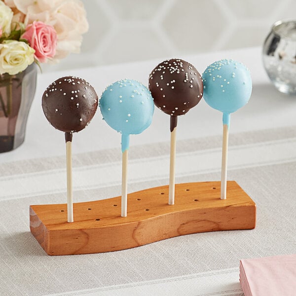 Four cake pops, two coated in chocolate and two in blue icing with white sprinkles, are displayed upright on a wooden holder with multiple holes.