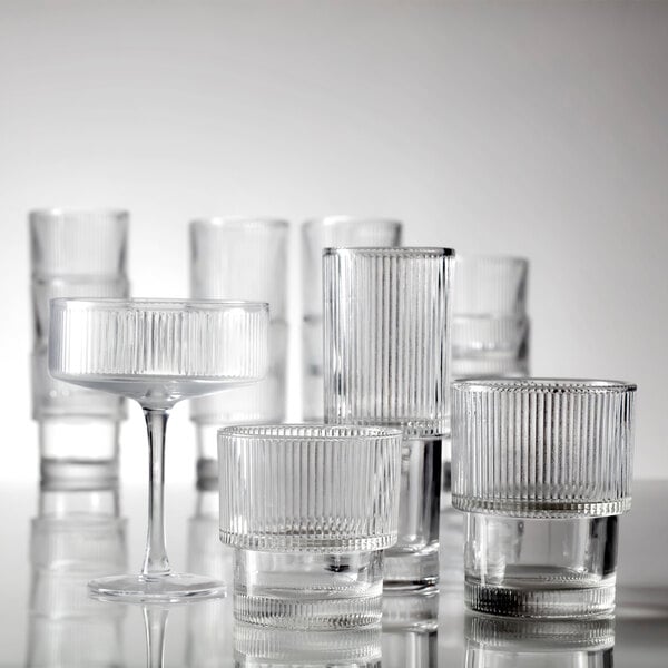 A group of clear, ribbed drinking glasses and a coupe glass are arranged on a reflective surface.