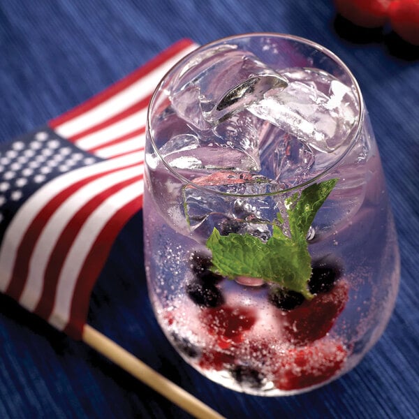A glass filled with ice cubes, berries, mint leaves, and a clear sparkling beverage sits next to a small American flag on a blue surface.