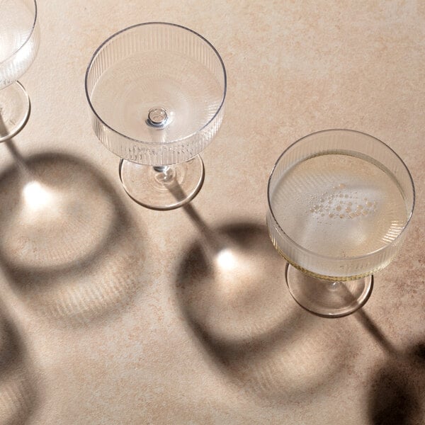 Three clear coupe glasses, one partially filled with a light-colored liquid, are casting distinct shadows on a beige surface.