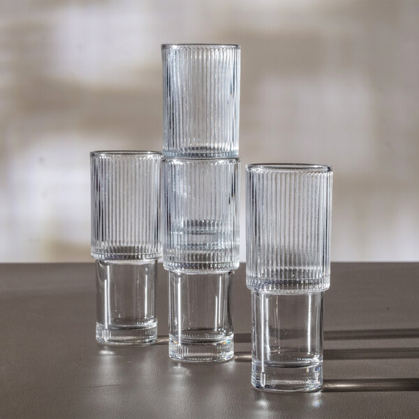 Four clear, vertically ribbed glasses are stacked and arranged on a flat surface with light and shadow in the background.