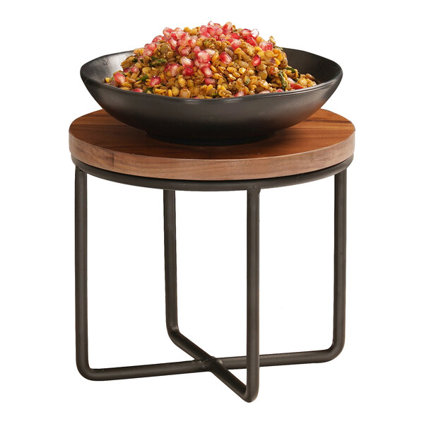 A small round table with a wooden top and black metal frame, holding a black bowl filled with a colorful food mixture.