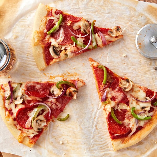 Three slices of pizza with pepperoni, mushrooms, green peppers, and red onions are arranged on parchment paper next to a pizza cutter and a jar of red pepper flakes.