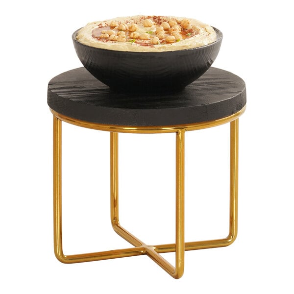 A small round black wooden platform with a gold metal frame, supporting a black bowl filled with hummus and garnished with chickpeas.