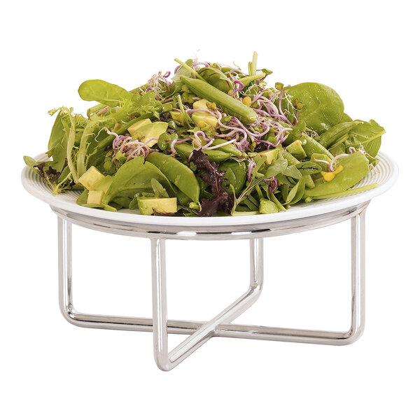 A white plate filled with salad is displayed on a round, chrome wire riser with a modern geometric design.