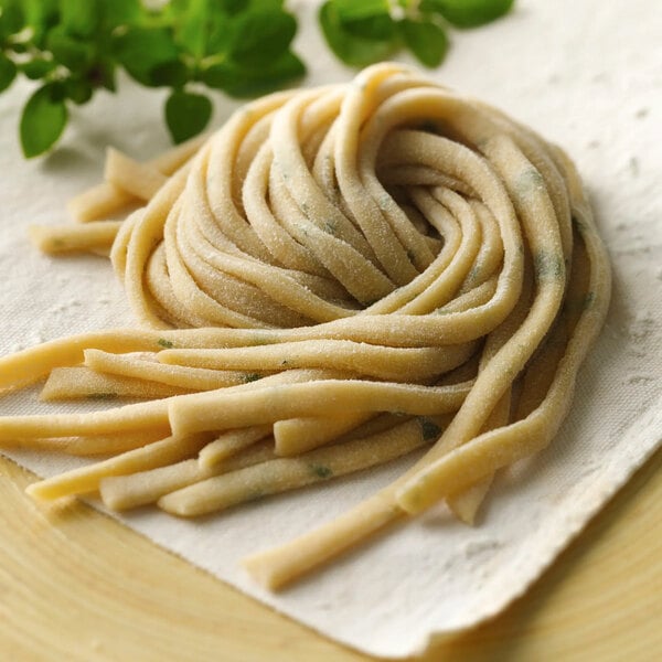 A nest of uncooked, flat, and slightly thick pasta noodles resting on a white cloth with green herbs in the background.