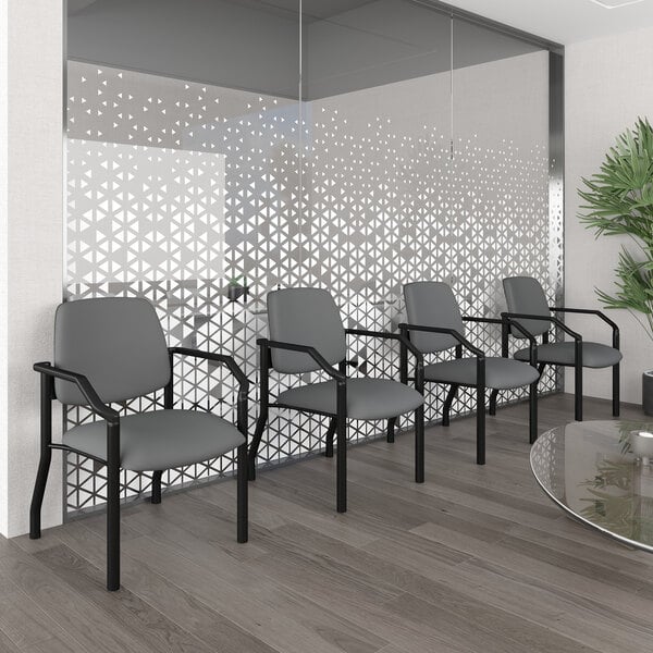 Four gray chairs with black metal frames and armrests are arranged in a row against a glass wall with a geometric pattern in a modern waiting area.