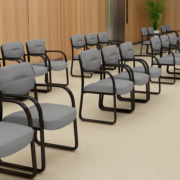 Several gray cushioned chairs with black sled bases and armrests are arranged in rows in a room with light wood paneling and beige flooring.