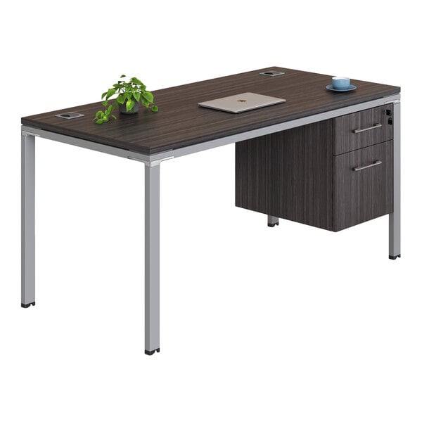 A modern office desk with a dark wood-like surface, metal legs, two drawers on one side, and items including a small plant, a closed laptop, and a cup with a saucer on top.