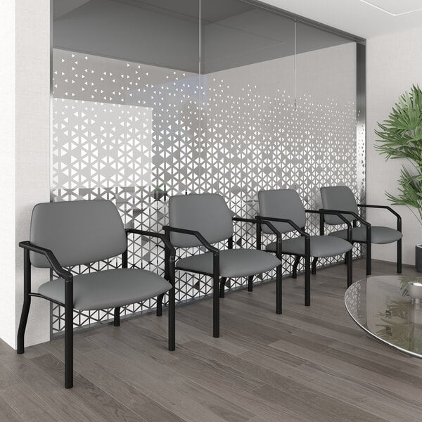 Four gray chairs with black metal frames and armrests are arranged in a row against a glass wall with a geometric pattern in a modern waiting area.