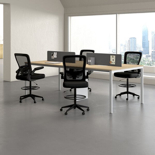 A modern office space with four black mesh drafting stools featuring flip-up arms and footrings, arranged around a shared desk with privacy dividers.