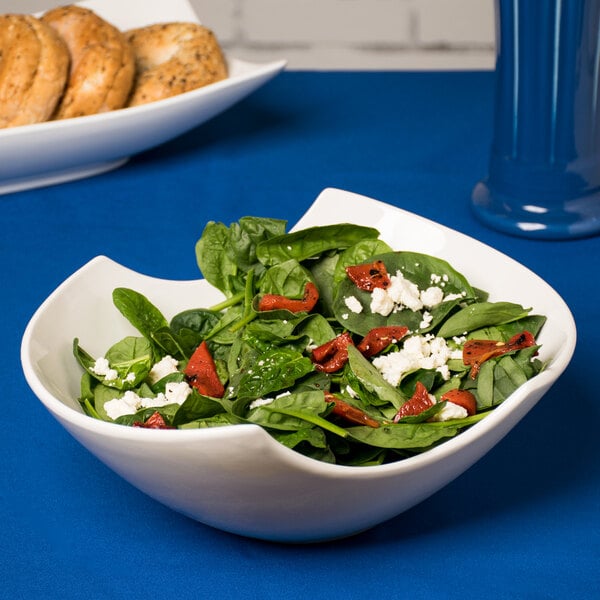 A white porcelain Samurai bowl filled with salad on a counter with bagels and a blue tablecloth.