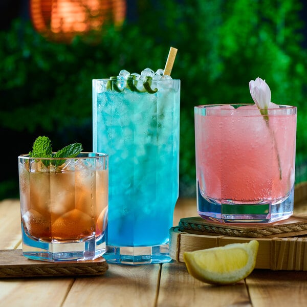 Three colorful drinks in clear glasses are displayed on a wooden surface, with garnishes including mint, a flower, and a lemon wedge, set against a blurred green background.