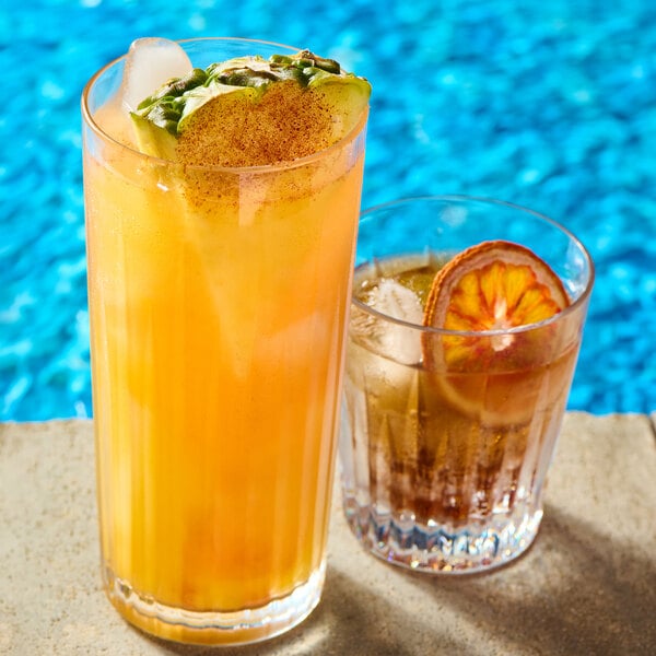 Two drinks are placed on a poolside surface: one is a tall, clear glass filled with an orange beverage, ice, and garnished with a pineapple slice, while the other is a shorter glass with a brown drink, ice, and a dried citrus slice, with a blue pool in th