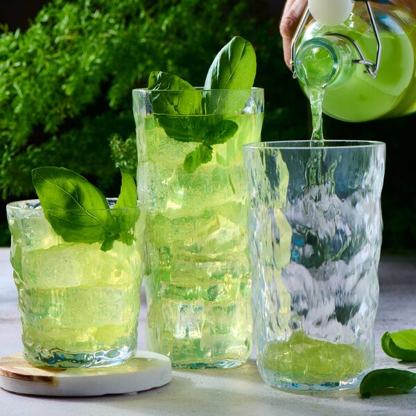 Three textured clear glasses filled with a green beverage, ice, and garnished with fresh basil leaves, with one glass being poured from a bottle.