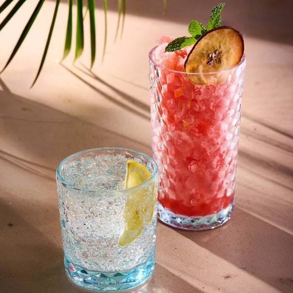 Two textured, light blue-tinted glasses are shown: one short glass with a clear, bubbly drink and a lemon wedge, and one tall glass with a red crushed ice drink garnished with a dried fruit slice and mint.