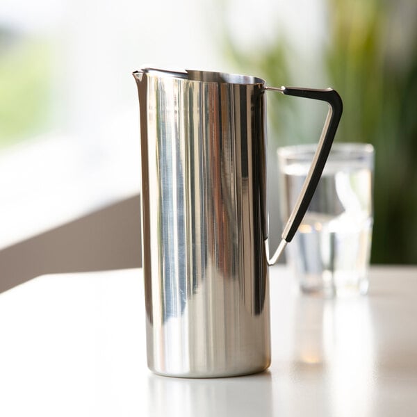 A tall, polished stainless steel pitcher with a sleek handle is placed on a white surface, with a glass of water in the background.