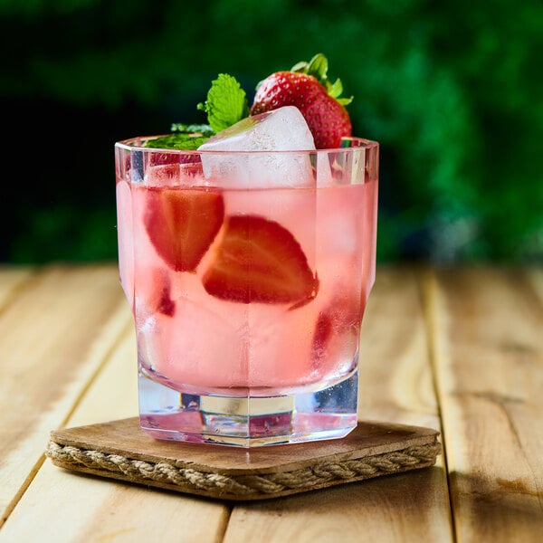 A clear glass filled with a pink beverage, ice cubes, strawberry slices, and garnished with a whole strawberry and mint, placed on a wooden coaster.