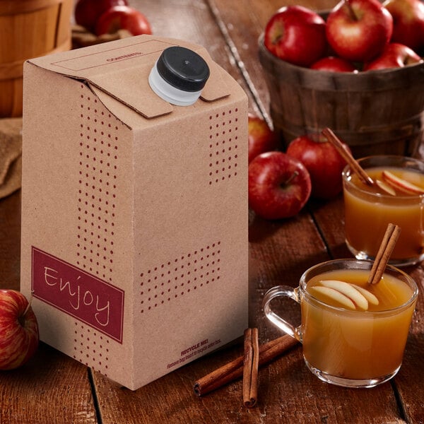 A cardboard beverage container with a plastic cap is placed on a wooden table next to glass mugs of apple cider garnished with cinnamon sticks and apple slices, with fresh apples in the background.