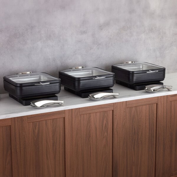 Three square black chafing dishes with glass lids and silver serving utensils are arranged on a marble countertop above wooden cabinets.