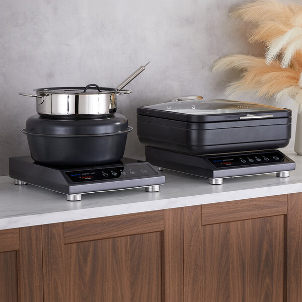 Two black induction cookers are placed on a marble countertop, one with a stainless steel pot and the other with a square black container with a glass lid.