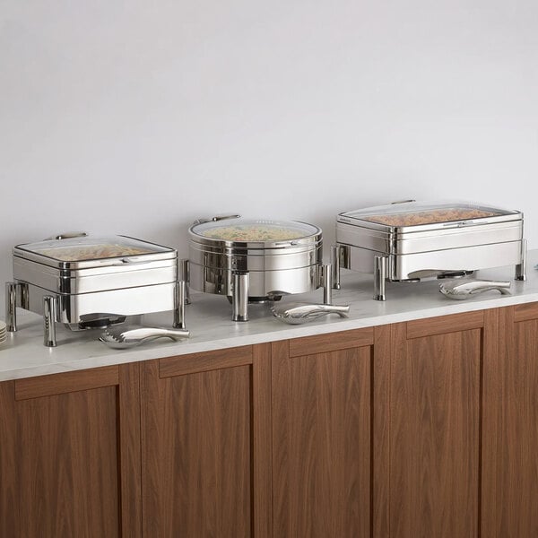 Three stainless steel chafing dishes with glass lids are displayed on a countertop, each with serving utensils placed in front.