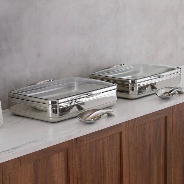 Two rectangular stainless steel buffet chafers with glass lids are placed side by side on a marble countertop, each accompanied by a serving utensil.