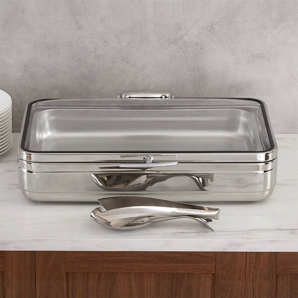 A rectangular stainless steel buffet chafer with a glass lid and a pair of serving tongs placed in front of it.