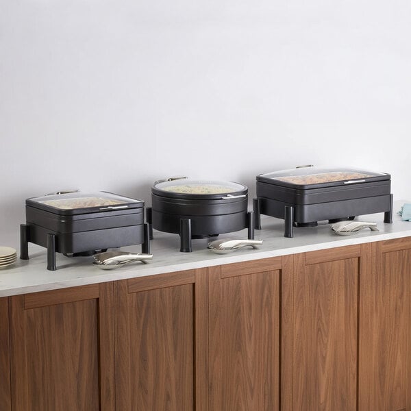 Three black buffet chafer stands with lids are arranged on a countertop above wooden cabinets, each accompanied by serving utensils.