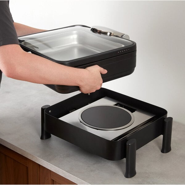 A person is placing a rectangular metal container with a glass lid onto a black induction buffet chafer stand on a countertop.