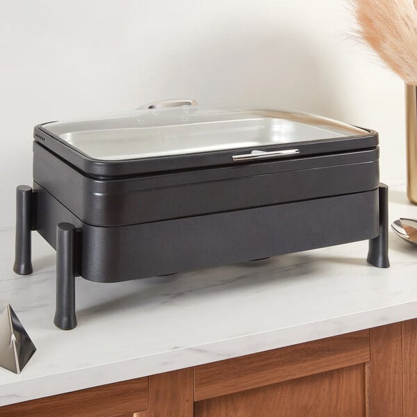 A rectangular black container with a glass lid and four short legs, placed on a countertop.