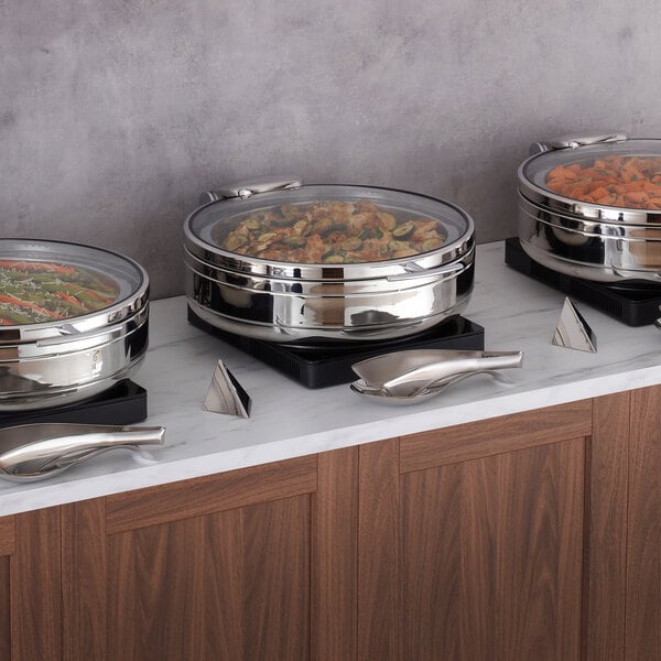 Three round stainless steel chafing dishes with glass lids are displayed on a marble countertop, each containing different types of food.