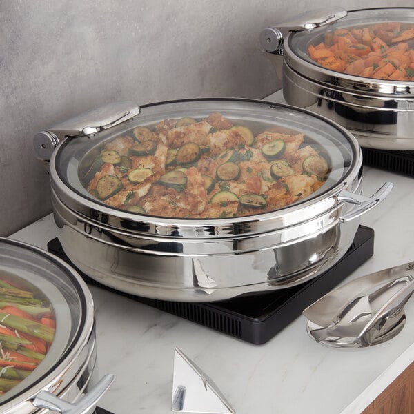 A round stainless steel chafing dish with a glass lid containing a cooked dish, placed on a countertop with serving tongs nearby.