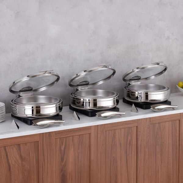 Three round stainless steel chafing dishes with glass lids are displayed on a countertop, each accompanied by a serving spoon and a small label holder.