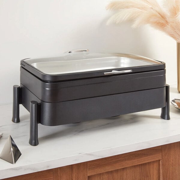 A rectangular black buffet chafer with a glass lid, elevated on four cylindrical legs, placed on a marble countertop.