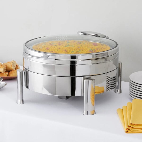 A round, stainless steel buffet server with a glass lid, containing yellow rice, is displayed on a white tablecloth next to plates, napkins, and bread.