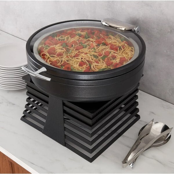 A round black chafing dish with a glass lid containing pasta, placed on a modern black stand on a countertop next to a stack of plates and serving utensils.