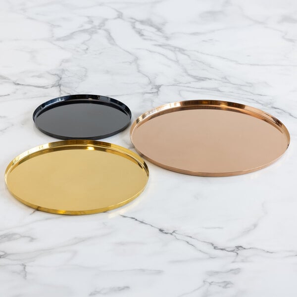 Three round metal trays in gold, black, and copper colors are displayed on a white marble surface.