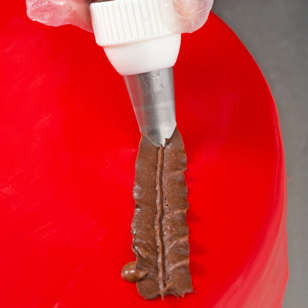 A piping bag fitted with a metal leaf tip is being used to pipe a strip of chocolate frosting onto a red surface.
