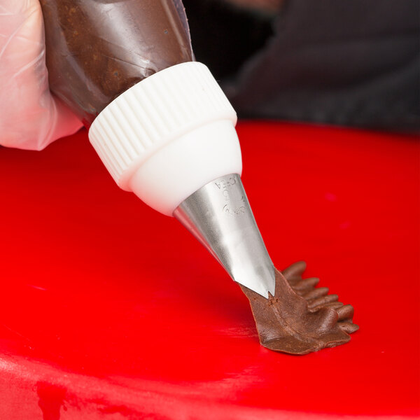 A close-up of a piping bag with a metal leaf tip being used to pipe chocolate frosting onto a red surface.