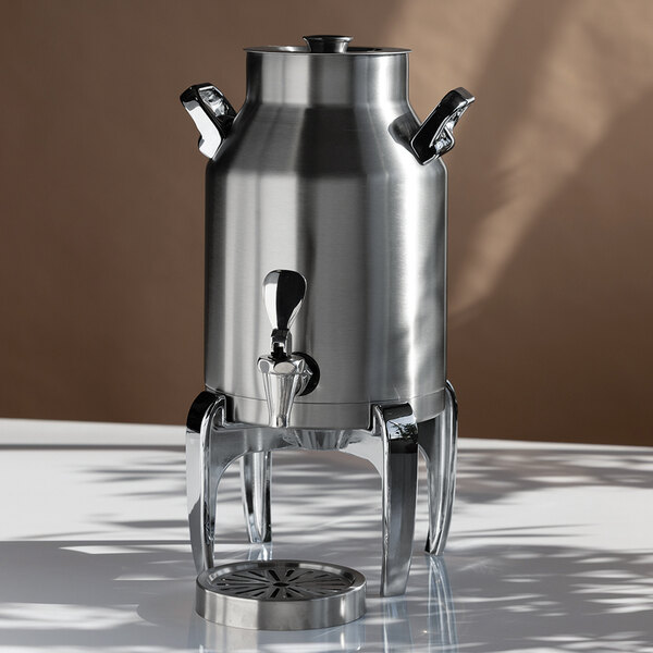 A stainless steel beverage urn with a spigot, two side handles, and a raised stand, placed on a reflective surface.