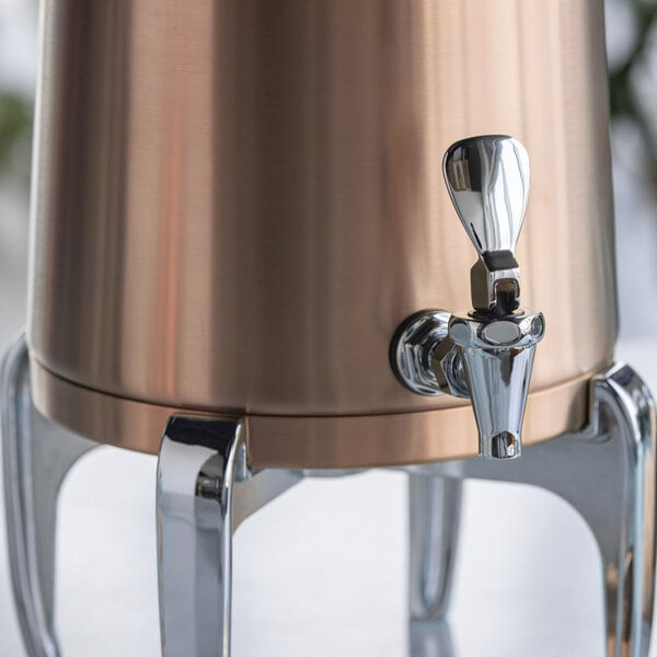 A close-up view of a rose gold and chrome beverage dispenser with a spigot and sturdy legs.